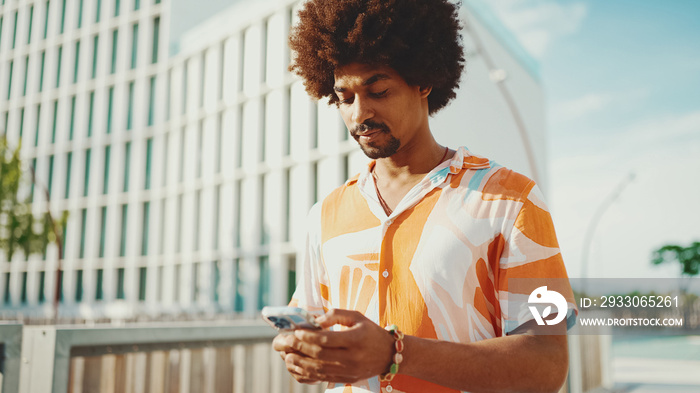 Closeup young African American man wearing shirt writes in social networks on mobile phone on urban street background. Camera moving forwards approaching to the person.