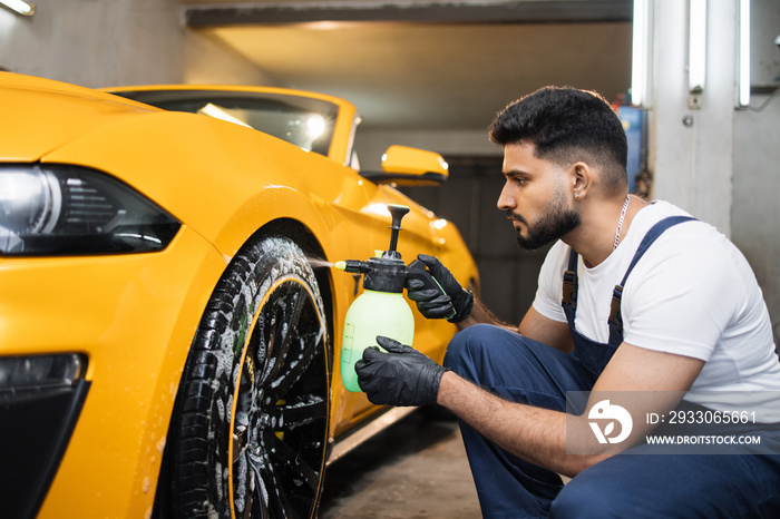 Washing a car in detailing service. Indoor shot of male worker in overalls, washing the car wheels rims with pressure water sprayer after cleaning it with special cleansing solution.