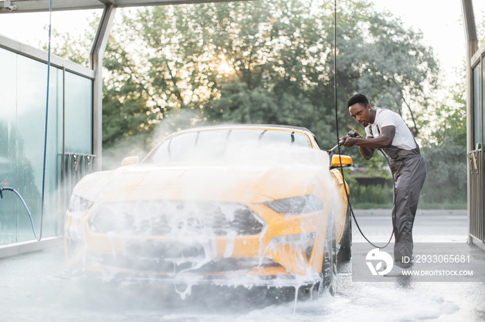 African stylish young focused man in work clothes washing his modern sport car with a water gun on self-service washing car station outdoors, rinsing the foam.