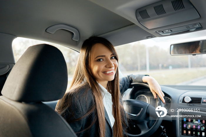 Driver turns to passenger seat and smiles at camera. Happy woman taxi driver sitting in car holding to steering wheel