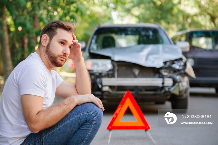 Portrait of upset man sitting on road near wrecked car after car accident with red triangle. Caucasian scary driver man sitting near his wrecked car on floor waiting for the insurance service.