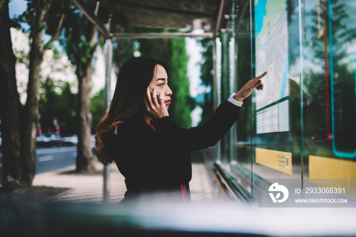 Pensive female tourist using mobile connection in roaming calling to friend while searching route of public transport on maps on bus stop, concentrated woman waiting for bus checking schedule