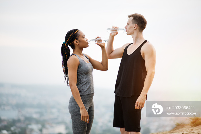 Side view of fitness young people in sport outfit drinking fresh cold water after outdoors activity. Multiracial couple leading a healthy and active lifestyles.