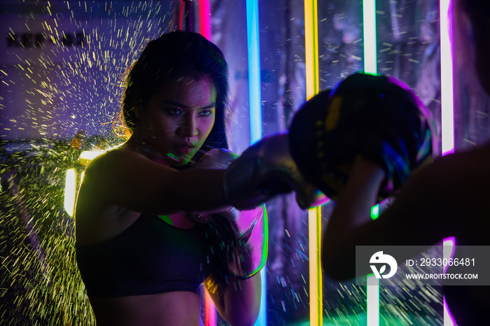 Beautiful 20s Asian Woman trains punching with coach on Silver gold Mitts Gloves. Office Girl exercise at Modern multi color Fashion Neon Muay Thai Boxing Gym with sweat water splash reflect light