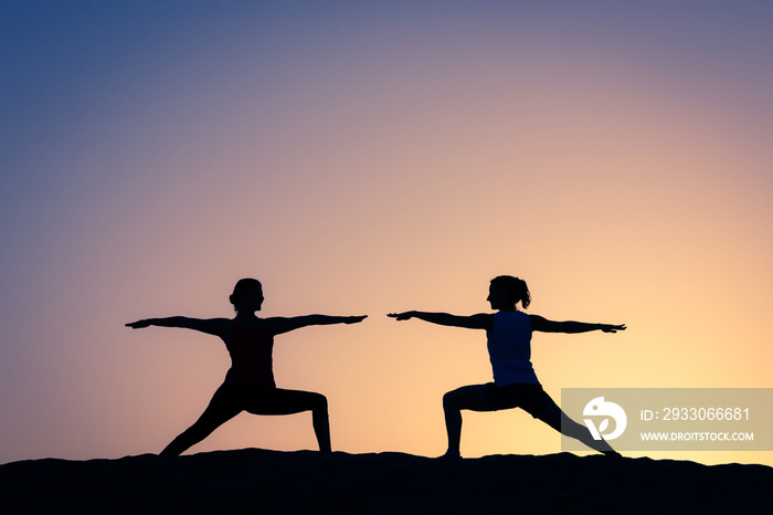 Women silhouettes practicing virabhadrasana two yoga pose at colorful sunset. Female yogis warrior asana at orange and blue twilight. Healthy lifestyle, outdoor workout concepts