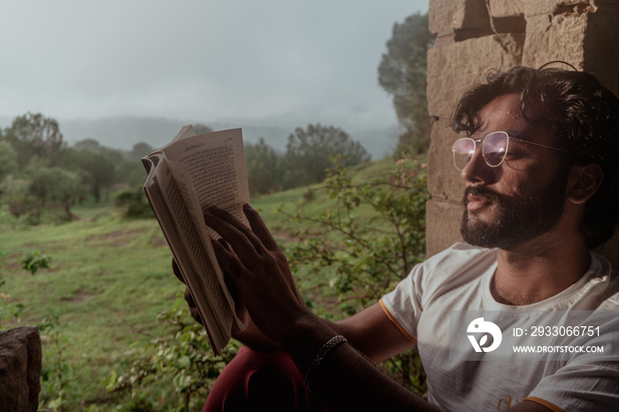 A hipster man wearing glasses flipping the pages of the book - reading book relaxing sitting near window view