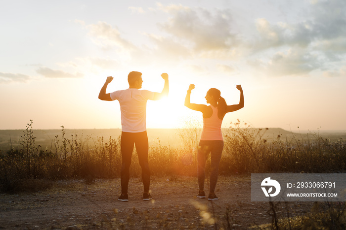 Couple of athletes flexing biceps towards the sunset. Couple of athletes celebrating running outdoor workout success.