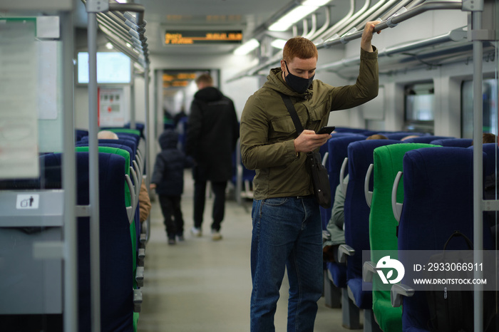 Handsome guy in respirator use a smartphone and keep social distancing to crowd while commuting in the metro or train. Infection and air pollution prevention concept