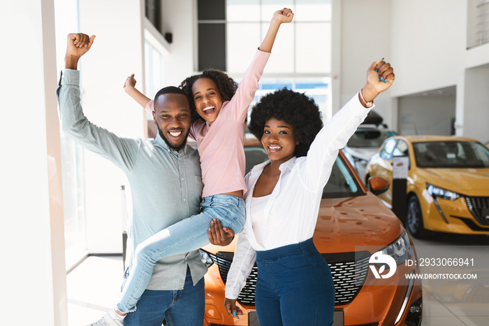 Excited black family of three lifting hands up, celebrating new car purchase in auto dealership