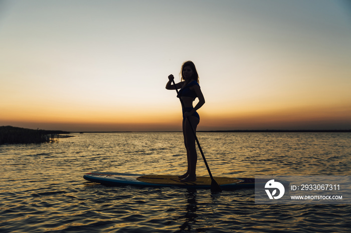 Young beautiful girl in a bikini is resting on a surfboard at sunset