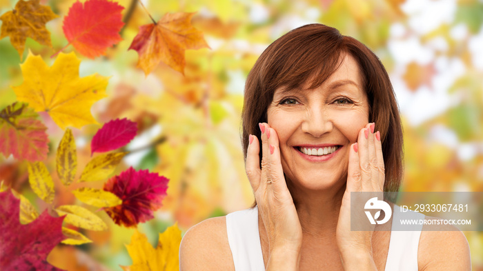 beauty, skin care and old people concept - portrait of smiling senior woman touching her face over autumn leaves and nature background