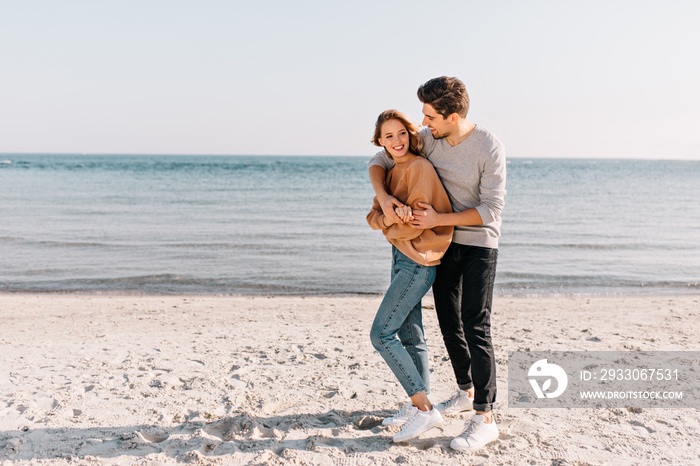 Ecstatic girl in jeans standing on sea background. Outdoor portrait of happy couple posing at sandy beach.