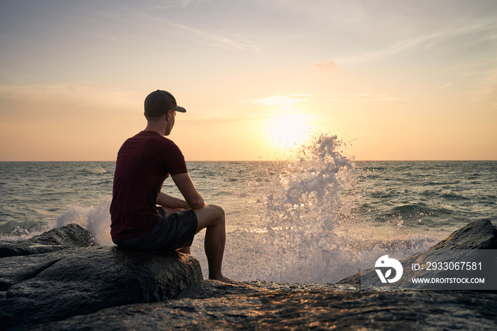 Rear view of man sitting on rock and watching beautiful sunset over ocean..