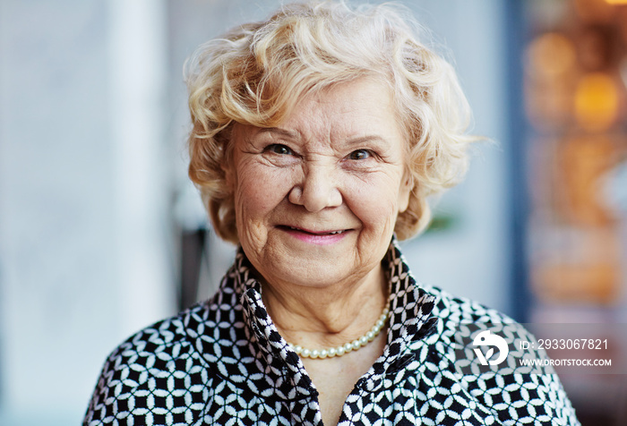 Head and shoulders portrait of cheerful senior woman in black-and-white cardigan and pearl necklace looking at camera with wide smile, blurred background