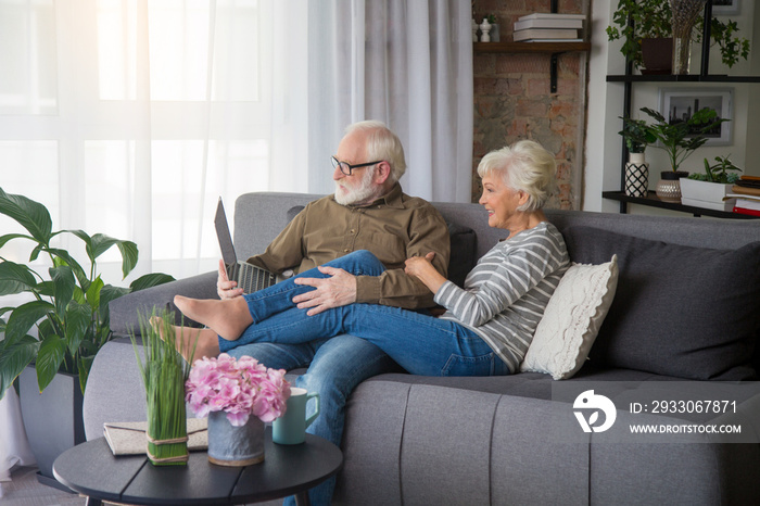 Family connection. Portrait of old couple looking into computer while talking. Lady is hugging husband while male is holding notebook sitting on couch at living room