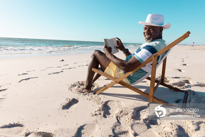 Smiling african american senior man wearing hat reading book on deckchair at beach under clear sky