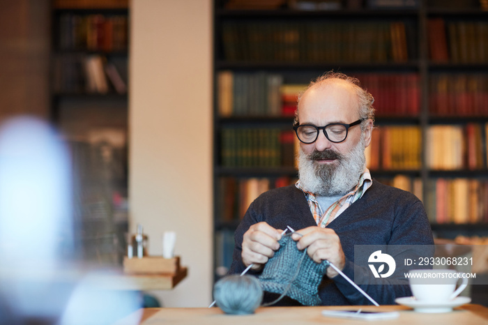 Bearded retired man in casualwear and eyeglasses knitting warm woolen winterwear