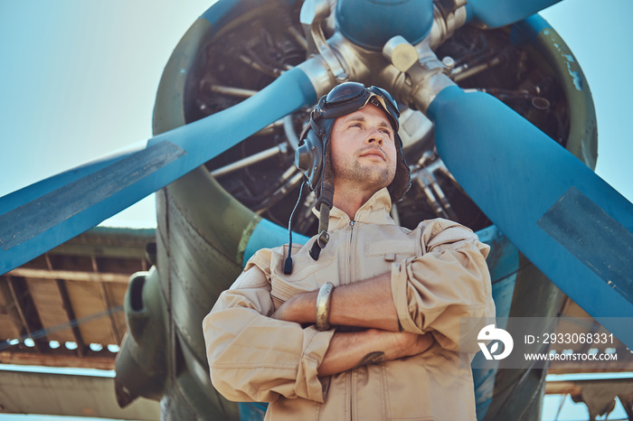 Valiant handsome pilot in a full flight gear standing with crossed arms near military airplane.