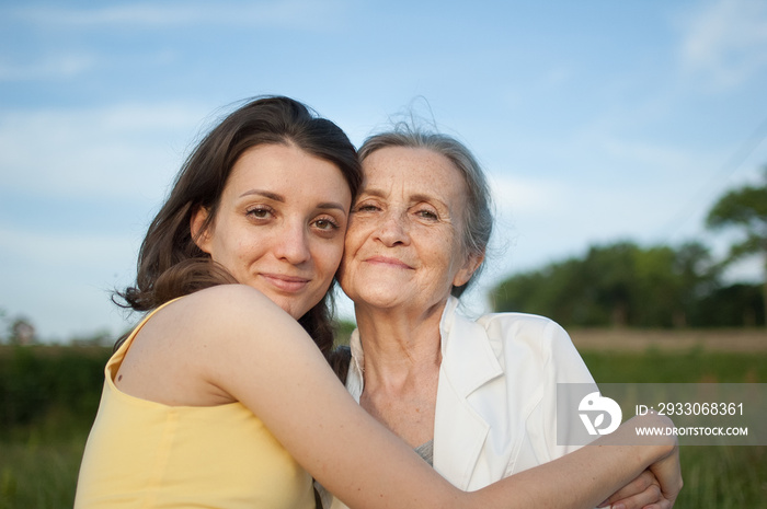 Senior mother with gray hair with her adult daughter looking at the camera in the garden and hugging each other during sunny day outdoors, mothers day