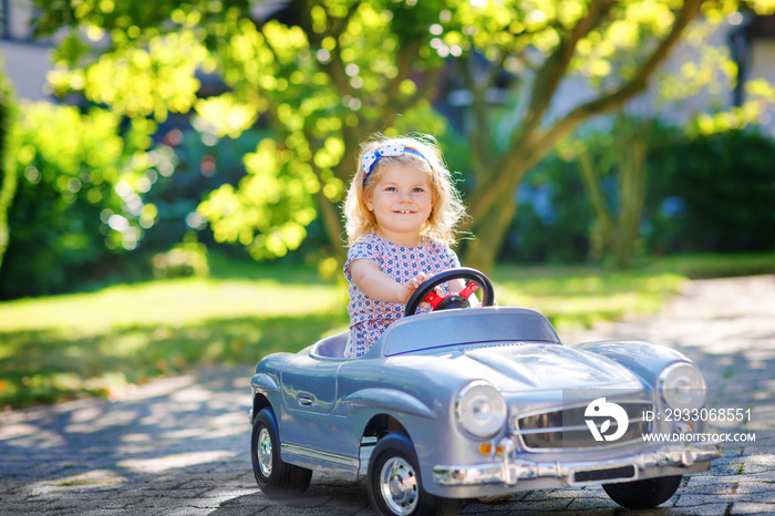 Little adorable toddler girl driving big vintage toy car and having fun with playing outdoors. Gorgeous happy healthy child enjoying warm summer day. Smiling stunning kid playing in domestic garden