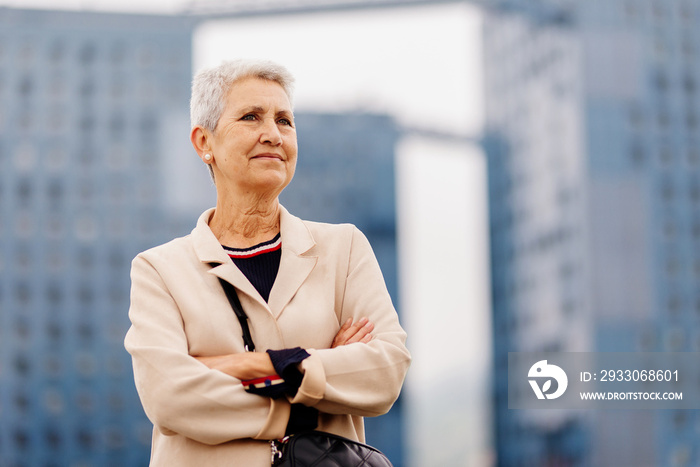 natural portrait of older woman with gray hair in , arms crossed and empowered in the city, smiling and looking at infinity.