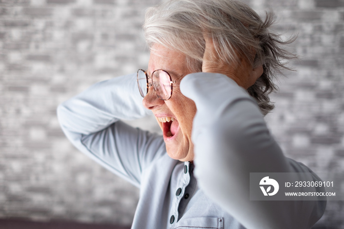 Desperate frustrated senior woman screaming in pain with hands on ears, sitting at home alone, senior woman suffering from divorce, bad relationship, mental illness