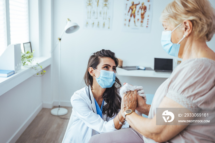 Elderly woman talking with a doctor while holding hands at home and wearing face protective mask. Worried senior woman talking to her general pratictioner visiting her at home during virus epidemic.