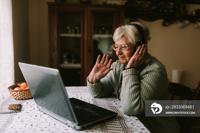 Smiling caucasian female senior with glasses and headphones on her head while sitting at a table in front of a laptop and waving to family during quarantine COVID - 19 coronavirus