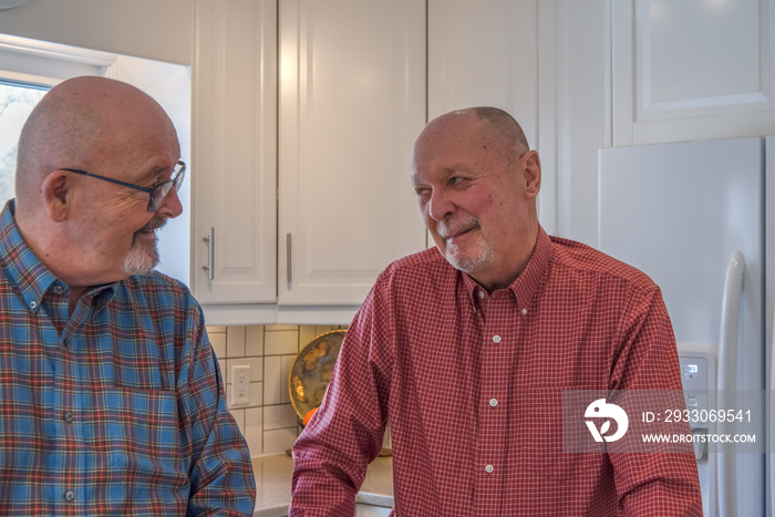 An elderly male gay couple in their kitchen smiling at each other while having a conversation.