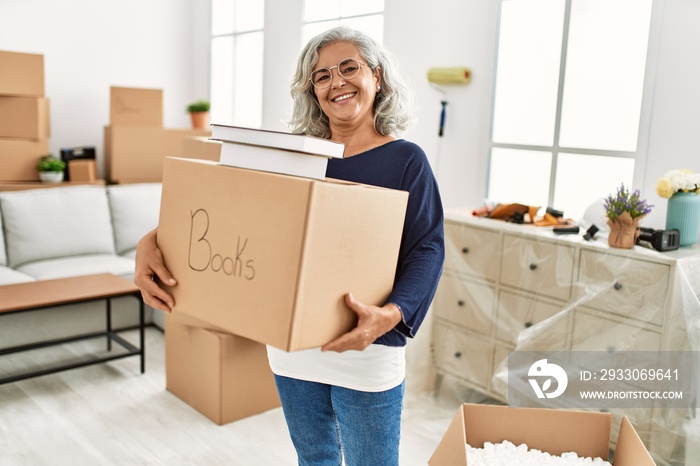 Middle age grey-haired woman smiling happy holding books cardboard box at new home.