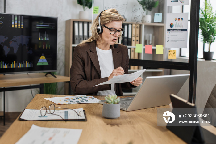 Caucasian aged business woman using modern headset and laptop for video conference at office. Attractive woman in formal wear having online meeting for reporting financial situation of company.