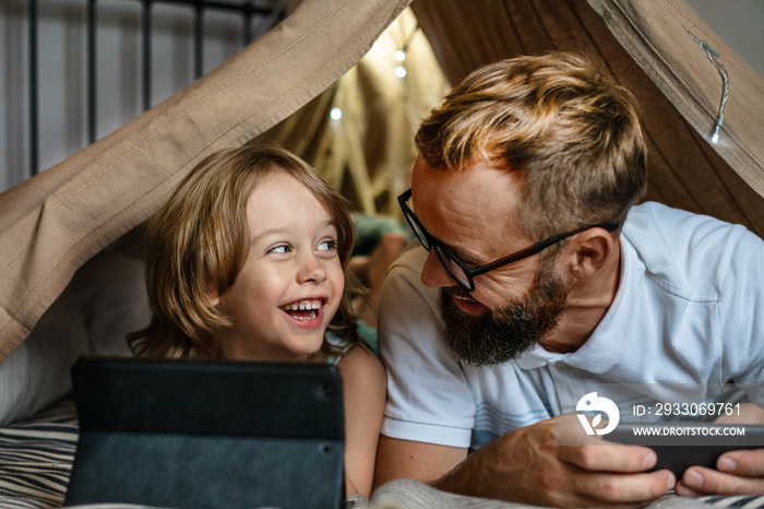 Portrait of a 6 year old boy and his father having fun playing in teepee tent. Father and son using digital tablet watching cartoons or playing computer games lying in kid tent at home.