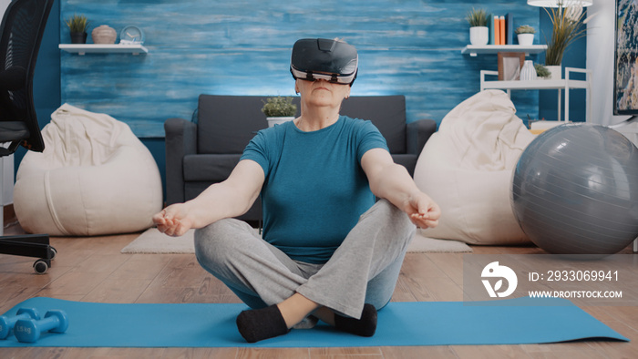 Aged woman using vr glasses to meditate on yoga mat. Elder person wearing virtual reality goggles while doing meditation in lotus position to relax before training. Calm adult with headset