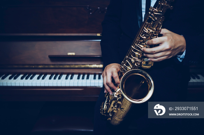 close up of Young Saxophone Player hands holding alto sax musical instrument with piano background in dark room, vintage tone, can be used for music background, copy space