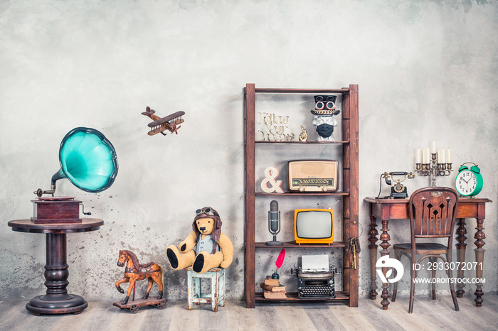 Antiques still life with old gramophone, Teddy bear, rocking horse and collection of outdated media, news makers and writers devices front concrete wall background. Vintage style filtered photo