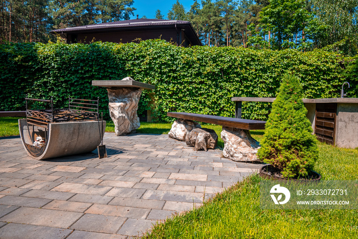 Beautiful sunny backyard with iron fire pit and benches on stone pavement, Garden rest area with fire pit surrounded by green shrubs