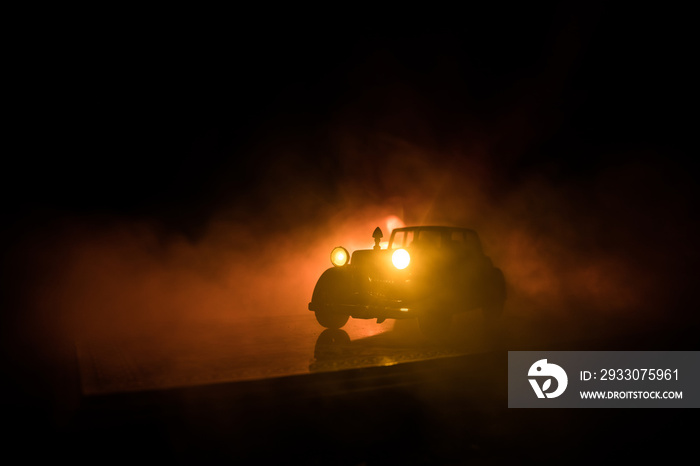 Silhouette of old vintage car in dark foggy toned background with glowing lights in low light.