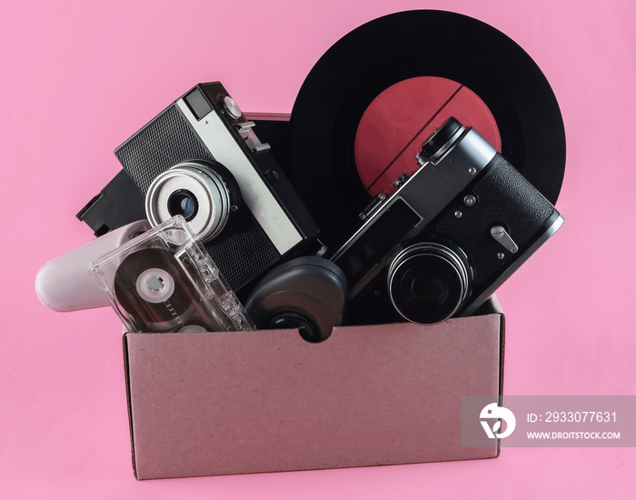 Cardboard box with retro film camera, gamepad,  vinyl record, phone tube, audio cassette on pink background. Antiques, trash