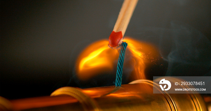 MACRO: Burning match lights up the short blue fuse of a miniature brass cannon.