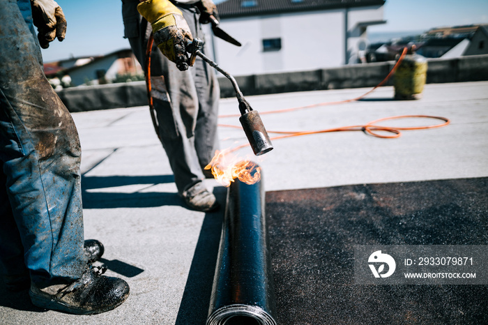 Construction workers insulating rooftop with bituminous membrane. Waterproofing details at construction site. Bituminous membrane waterproofing system details and installation on flat rooftop.