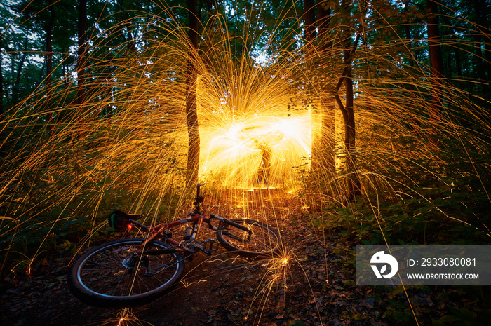 Burning steel wool spinned in the forest with a bicycle in the foreground . Showers of glowing sparks from spinning steel wool