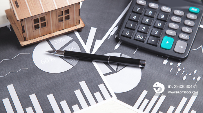 Wooden house model with a calculator and pen on financial graphs.