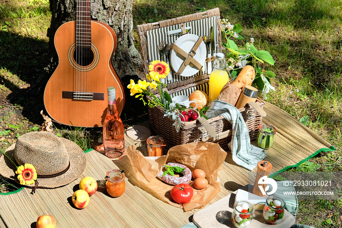 Vintage picnic basket, hamper with baguette and lemonade outdoors on a grass with cheese, mozzarella, tomatoes, cherries, vine. Guitar, snacks on straw mat. Eco friendly picnic outdoors.