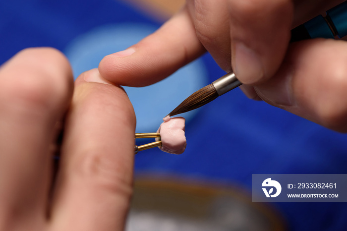 Dental technician applies ceramic material to the crown of a dental implant in a dental laboratory using tweezers and a brush. Dental technology close-up.