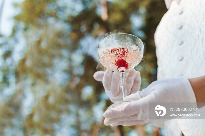 Waiter in white gloves gives wine glass with shampagne, red cherry, and white smoke of dry ice and gives to customer. Catering for wedding ceremony and business meeting. Empy left side for text.