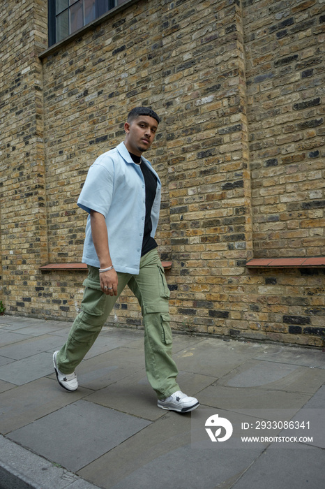 Man wearing unbuttoned shirt and cargo pants walking along brick wall