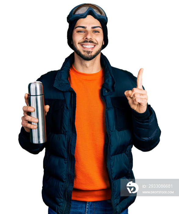 Young hispanic man with beard wearing snow wear and sky glasses holding thermo smiling with an idea or question pointing finger with happy face, number one