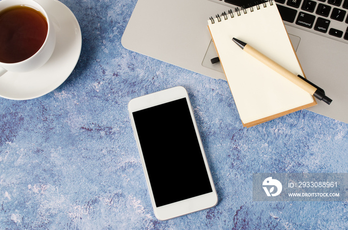 White smartphone with black blank screen on office desk with laptop, empty notebook and cup of tea. Mock up of phone.