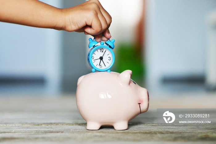 Save time concept with child’s hand holding alarm clock over a ceramic piggy bank