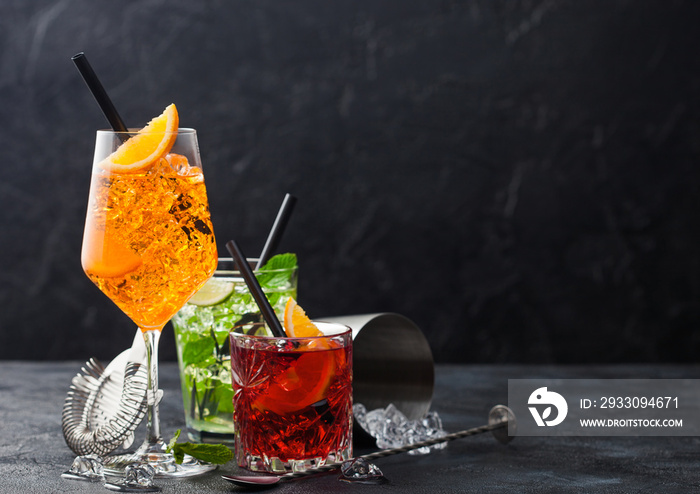 Glasses of spritz,mojito and negroni cocktails with ice cubes and lime and orange slices with mint leaf and black straw on black background with strainer and shaker.